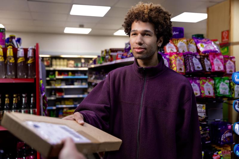Person in a purple jacket receives a package in a convenience store, surrounded by colourful snacks and drinks on shelves.