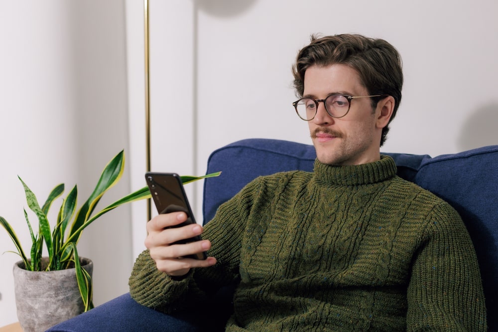 Man with glasses and mustache, wearing a green sweater, sitting on a blue chair, looking at a smartphone. A potted plant is beside him.
